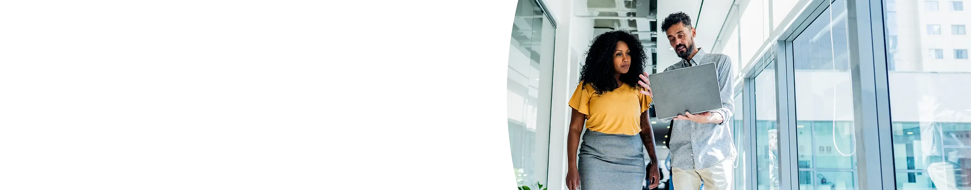 A man and woman walking down a bright, modern office hallway while looking at a laptop and discussing work.